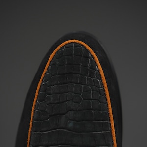 May include: Close-up of a black shoe toe with a textured, crocodile-like pattern. The shoe has an orange trim around the toe area. The shoe is made of a dark material, possibly suede or leather.