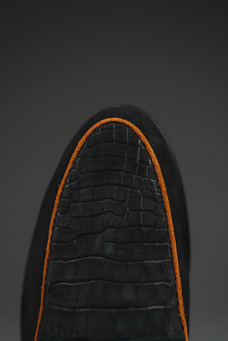May include: Close-up of a black shoe toe with a textured, crocodile-like pattern. The shoe has an orange trim around the toe area. The shoe is made of a dark material, possibly suede or leather.