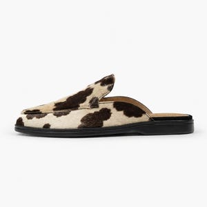 May include: A slip-on mule shoe featuring a cow print pattern with dark brown and cream-colored fur texture. The footwear has a pointed toe, a low-profile silhouette, and a solid black flat sole with a matching black interior lining.