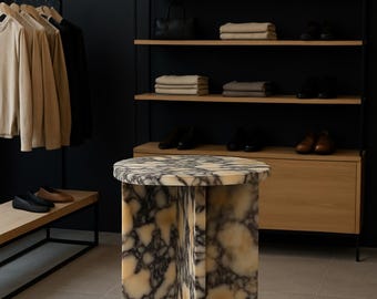Puede incluir: Una mesa auxiliar de mármol con tapa redonda y un diseño de base único. La mesa presenta un patrón veteado crema y marrón. El fondo incluye una barra de ropa, estantes con bolsos y zapatos.