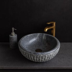 Peut inclure: Un lavabo en pierre grise avec un extérieur texturé, associé à un robinet doré et un distributeur de savon assorti. L'évier rond repose sur un comptoir sombre, créant une esthétique de salle de bain moderne.