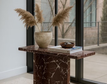 May include: A modern side table with a red marble top and base. A beige ceramic vase with dried plants sits on the table, along with a book and a small wooden bowl. The table is placed near a large window.