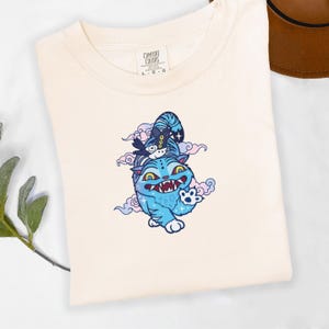 May include: Cream-colored t-shirt featuring a blue cartoon cat with a mischievous expression, set against a backdrop of pink and blue clouds. The cat has sharp teeth and is adorned with a hat. The shirt has a Comfort Colors tag.