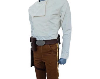Pode incluir: Um traje com uma camisa creme de manga comprida com gola assimétrica, combinada com calças castanhas. O traje inclui um cinto castanho com fivela prateada, uma coldre e detalhes dourados nas calças. O traje é exibido em um manequim.