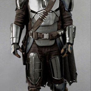 May include: A full-length image of a Mandalorian warrior in silver and dark armour. The helmet is polished silver, and the chest plate is a mix of silver and grey. The warrior wears a brown belt with ammunition pouches and a dark cape.
