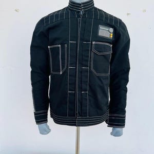 Puede incluir: Chaqueta negra con costuras blancas. Tiene cuello alto, dos bolsillos en el pecho y un parche en el brazo. La chaqueta se exhibe en un maniquí.