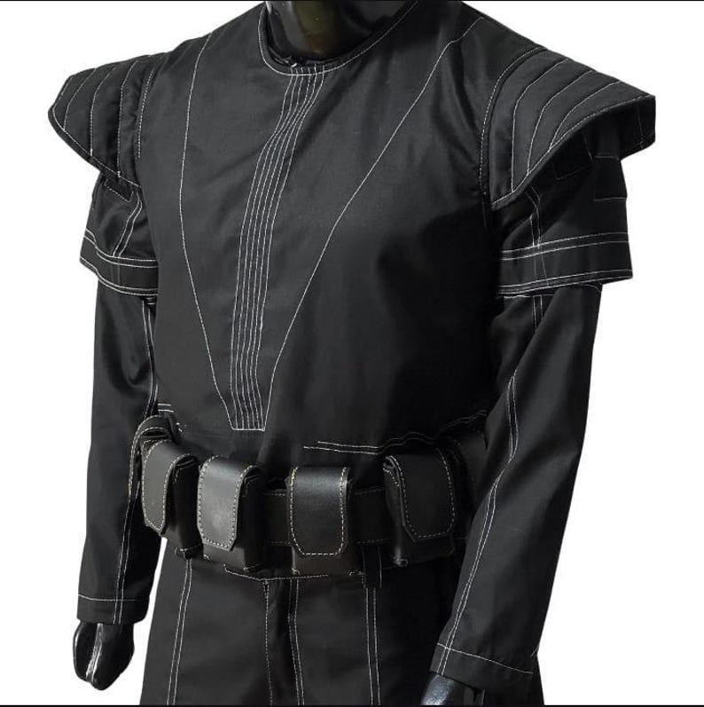 May include: Black costume with white stitching details. The outfit features padded shoulder accents and a belt with multiple pouches. The long-sleeved garment has a high neckline and is made of a matte fabric.