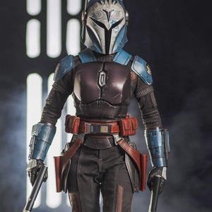 May include: A detailed action figure of a character in a blue and silver helmet and armor, holding two blasters. The figure has a red belt with pouches and is posed against a dark background.