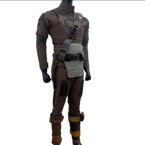 May include: A brown and grey costume with a high collar, shoulder pads, and a utility belt. The costume includes a chest piece, a shoulder strap with pouches, and brown trousers with knee pads. The mannequin is wearing black gloves and boots.