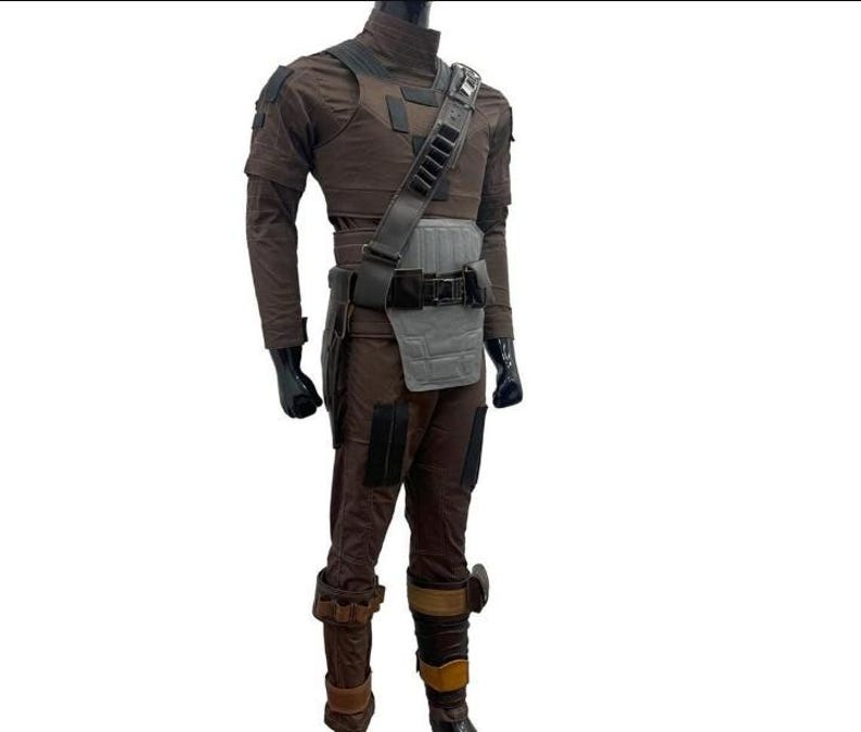 May include: A brown and grey costume with a high collar, shoulder pads, and a utility belt. The costume includes a chest piece, a shoulder strap with pouches, and brown trousers with knee pads. The mannequin is wearing black gloves and boots.