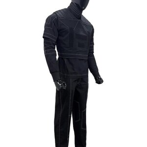 May include: A black long-sleeved shirt and matching trousers set displayed on a mannequin. The outfit features a high collar, structured shoulder panels, and prominent white contrast stitching throughout the garment, creating geometric patterns.