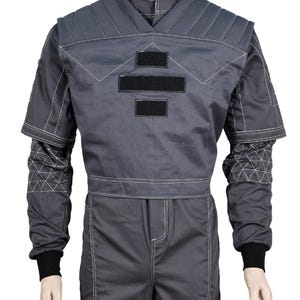 May include: A gray jumpsuit with white stitching and black accents. The top features a V-neck design, padded shoulders, and three black rectangular patches. The sleeves have geometric patterns near the cuffs, and the cuffs are black.