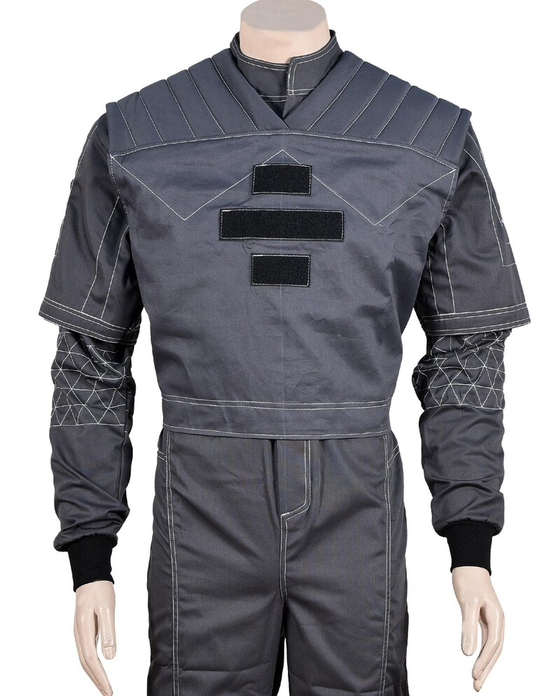 May include: A gray jumpsuit with white stitching and black accents. The top features a V-neck design, padded shoulders, and three black rectangular patches. The sleeves have geometric patterns near the cuffs, and the cuffs are black.
