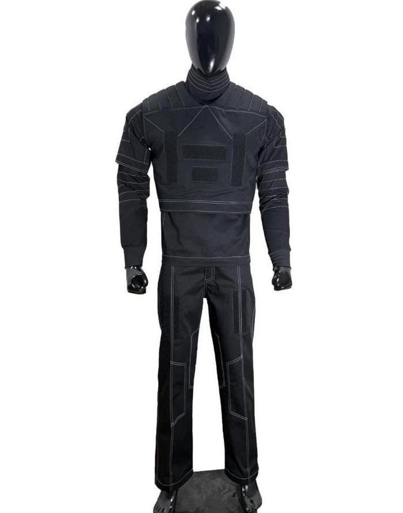 May include: A black tactical-style two-piece outfit consisting of a long-sleeved top with a high collar and matching trousers. The garment features prominent white contrast stitching, structured shoulder panels, and rectangular hook-and-loop patches.