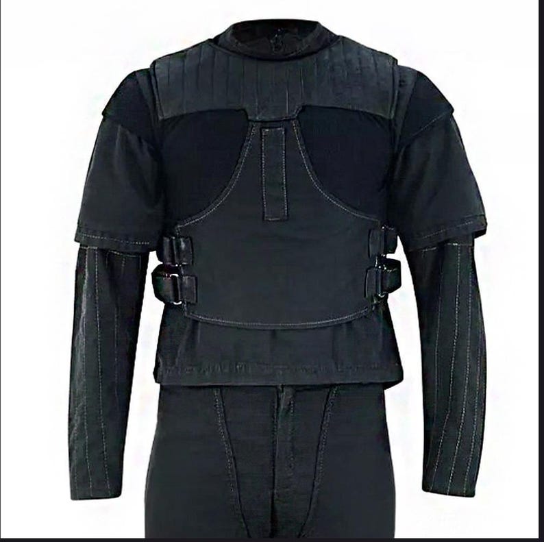 Peut inclure: Gilet tactique noir avec manches courtes et col montant. Le gilet pr&eacute;sente une section sup&eacute;rieure rembourr&eacute;e et un panneau central avec deux boucles. Les manches longues ont des coutures verticales. Le gilet est port&eacute; sur une chemise et un pantalon noirs.
