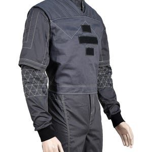 May include: A gray jumpsuit with white stitching details. The jacket has a stand-up collar, black patches, and geometric patterns on the sleeves. The pants have a straight leg design. The cuffs are black.