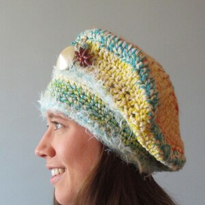 Peut inclure: Un bonnet en crochet de couleur jaune, bleu et orange. Le bonnet est orné d'une bordure blanche moelleuse et d'une perle et d'une fleur rouge.