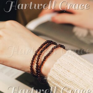 Peut inclure: Trois bracelets de perles rouge foncé ornent un poignet. Les bracelets sont composés de petites perles rondes. Une manche de pull crème est en arrière-plan. Le texte "Hartwell Grace" est visible.