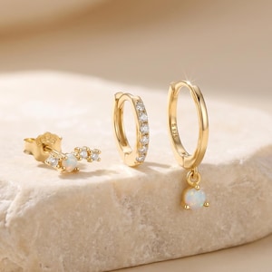 Puede incluir: Un conjunto de pendientes dorados expuestos sobre una piedra clara. El conjunto incluye un pendiente de botón con un diseño curvo, un pendiente de aro pequeño con piedras transparentes y un pendiente de aro con una piedra colgante similar al ópalo. Los pendientes están sobre un fondo suave y neutro.