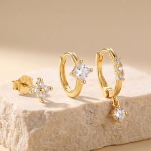May include: A set of three gold earrings. One is a stud with a flower design, another is a small hoop with a square-cut clear stone, and the third is a hoop with multiple small clear stones and a dangling clear stone.