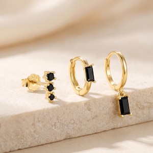 Puede incluir: Tres pares de pendientes dorados con piedras negras. Un par presenta un diseño de tachuelas con tres piedras redondas. El segundo par es un aro pequeño con una piedra rectangular. El tercer par tiene un aro con una piedra rectangular colgante.