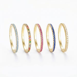 Könnte beinhalten: Fünf goldfarbene Ringe mit Bändern aus farbigen Edelsteinen. Die Ringe sind in einer Reihe angeordnet, jeder mit einer anderen Edelsteinfarbe: hellblau, Regenbogen, rosa, dunkelblau und klar. Der Hintergrund ist weiß.