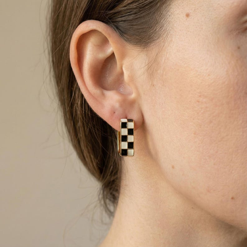 K&ouml;nnte beinhalten: Goldfarbene, rechteckige Creolen mit einem schwarz-wei&szlig;en Schachbrettmuster. Die Ohrringe sind ein auff&auml;lliges Accessoire mit einem klassischen Design und einem modernen Touch. Die schwarzen und wei&szlig;en Quadrate erzeugen einen auff&auml;lligen Kontrast.