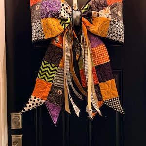 May include: A large quilted fabric bow featuring a patchwork design of orange, purple, black, and white fabrics with various patterns including skulls, pumpkins, and chevrons. The bow has long, dangling fabric ribbons and is hung on a black door.