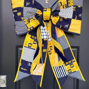 Puede incluir: Un gran lazo decorativo hecho de tela acolchada con el logotipo y los colores de la Universidad de Michigan. El diseño incluye patrones en azul marino, amarillo y blanco con lunares, rayas y el símbolo de la M en bloque por toda la tela.