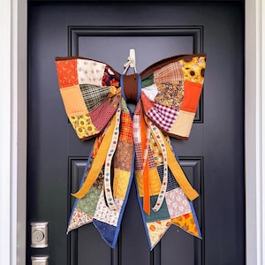 May include: A large decorative bow made of quilted fabric patches in various autumn-inspired patterns and colors, including orange, brown, yellow, and green. The bow features sunflower prints, plaid, and leaf motifs with long, trailing fabric tails.