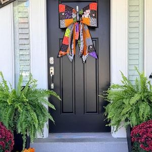 Può includere: Un grande fiocco decorativo realizzato in tessuto patchwork con vari motivi a tema Halloween in arancione, nero, viola e bianco. Il fiocco presenta lunghi nastri fluenti con stampe diverse, tra cui pois, righe e motivi stagionali.