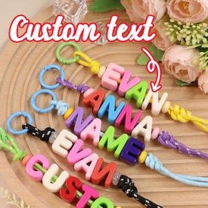 Op de afbeelding: Kleurrijke sleutelhangers met namen als "EVAN" en "ANNA" van kralen. Elke sleutelhanger heeft een ander kleurenschema voor de letters en koorden. De tekst "Custom text" is zichtbaar in de linkerbovenhoek.