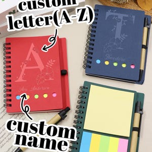 Pode incluir: Uma coleção de cadernos personalizados em vermelho, azul marinho e verde. Cada caderno apresenta uma letra ou nome personalizado, com um design floral. Os cadernos incluem uma caneta e notas adesivas. O texto "custom letter (A-Z)" e "custom name" são visíveis.