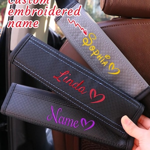 May include: Custom embroidered seat belt covers in gray, black, and dark gray. Each cover features a personalized name in a different color, such as yellow, red, and purple, with a heart symbol. The image also shows spools of thread in various colors.