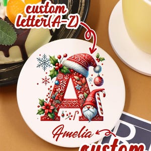 Può includere: Un sottobicchiere in ceramica bianca con una lettera "A" rossa decorata con elementi natalizi, tra cui un cappello di Babbo Natale, agrifoglio e ornamenti. Il nome "Amelia" è scritto sotto. Il testo sull'immagine dice "custom letter (A-Z)" e "custom name".