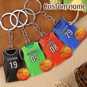 May include: Four basketball jersey keychains in black, green, red, and blue. Each keychain features a jersey design with a name and number, along with a basketball. The text "Custom name" is also visible.
