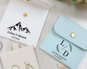 Könnte beinhalten: Eine Auswahl kleiner, personalisierter Geschenkbeutel in Weiß und Hellblau, mit goldenen Druckknöpfen. Der weiße Beutel zeigt eine Berg-Grafik und die Namen "Evelina & Maxime" mit einem Datum. Der hellblaue Beutel hat Initialen und ein Datum.