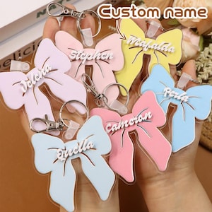 May include: A collection of personalized bow-shaped keychains in various pastel colors, including pink, yellow, and blue. Each keychain features a name in a contrasting white font, attached to a silver-colored clasp and ring. The text "Custom name" is visible at the top.
