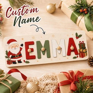 May include: A rectangular wooden name puzzle featuring the name EMMA in red, green, and white letters with small wooden knobs. The board is decorated with a cartoon Santa Claus, holly leaves, and stacked gift boxes on a light wood surface.