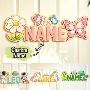 Puede incluir: Un letrero de pared de madera con el texto NAME en letras rosas, decorado con una flor blanca grande y mariposas rosas y azules. Debajo, tres ejemplos muestran el nombre LEO con astronautas, NAME con unicornios y NAME con dinosaurios.