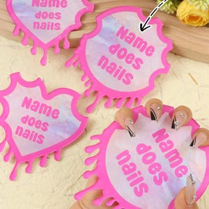 Pode incluir: Estênceis rosa para nail art com o texto "Name does nails" em fonte cursiva. Os estênceis têm um formato único com um design de gotejamento. A imagem também inclui o texto "Custom text".