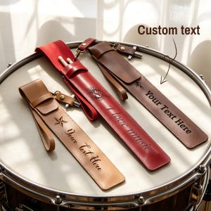 Op de afbeelding: Drie leren drumstokhouders in tan, rood en bruin, elk met een riem en sluiting. De rode houder heeft "I love music" gegraveerd, en de andere "Your Text Here". De houders liggen op een trommel.