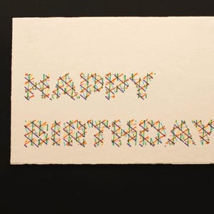 May include: A cream-coloured birthday card with the words "HAPPY BIRTHDAY" stitched in colourful thread. The letters are made of intersecting lines in various colours, including red, blue, green, and yellow.