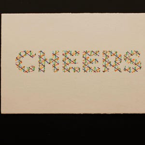 May include: A cream-coloured rectangular greeting card featuring the word "CHEERS" embroidered in a cross-stitch style. The letters are formed with colourful threads in red, yellow, green, blue, and orange.