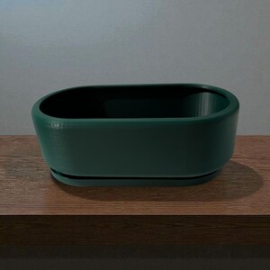 Puede incluir: Una jardinera ovalada de color verde oscuro con una base a juego, colocada sobre una superficie de madera. La jardinera tiene un acabado liso y mate y un interior profundo. Ideal para plantas peque&ntilde;as o suculentas.