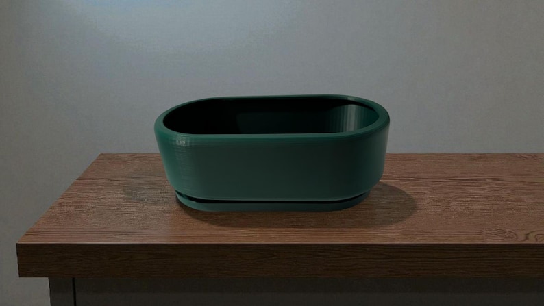 Puede incluir: Una jardinera ovalada de color verde oscuro con una base a juego, colocada sobre una superficie de madera. La jardinera tiene un acabado liso y mate y un interior profundo. Ideal para plantas peque&ntilde;as o suculentas.