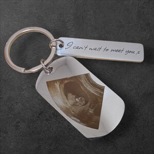 Op de afbeelding: Een zilveren sleutelhanger met twee rechthoekige labels. Eén label heeft een echografiebeeld, de andere heeft de gegraveerde tekst "I can't wait to meet you x". De sleutelhanger heeft een zilveren ring.