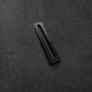 May include: A black metal rectangular object with a long, narrow, central cutout. The object is positioned diagonally against a dark, textured background, creating a contrast in tones and textures.