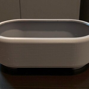 Puede incluir: Un recipiente rectangular blanco y gris con bordes redondeados. El recipiente tiene un interior gris y se asienta sobre una superficie oscura. El exterior tiene un aspecto texturizado y en capas.