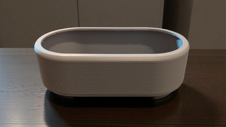 Puede incluir: Un recipiente rectangular blanco y gris con bordes redondeados. El recipiente tiene un interior gris y se asienta sobre una superficie oscura. El exterior tiene un aspecto texturizado y en capas.